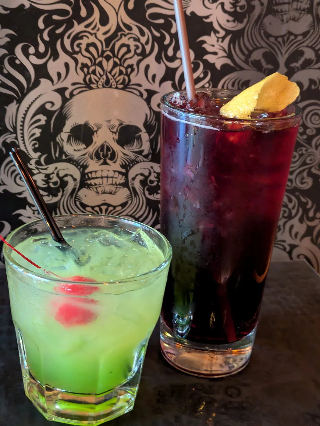 Two cocktails, one bright green and one deep purple, set against the bar's black and cream damask wallpaper