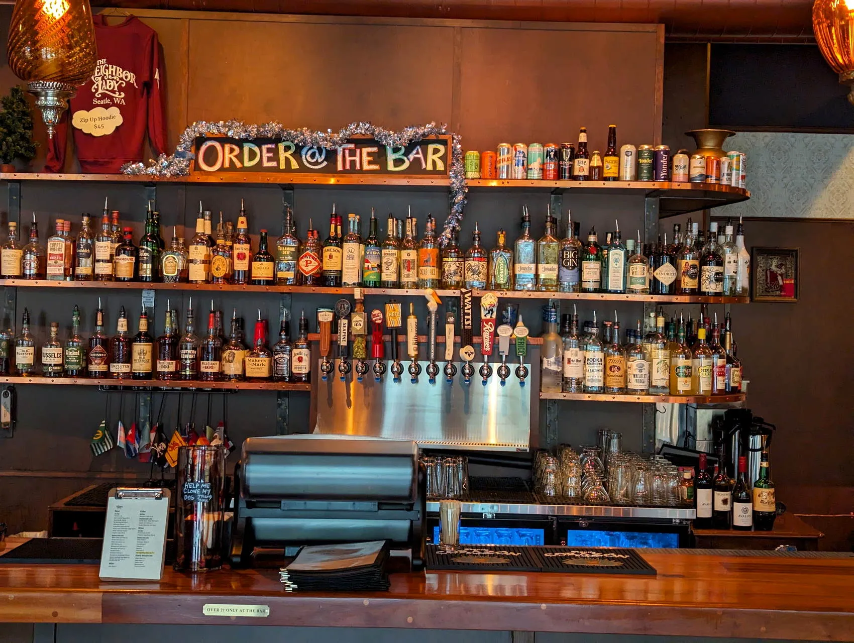 The back bar at The Neighbor Lady in Seattle, copper shelves stacked with whiskey and a hand-painted Order at the Bar sign