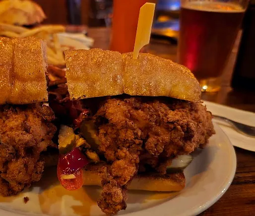 Fried chicken sandwich with pickles and hot sauce on a soft bun, a pint behind it